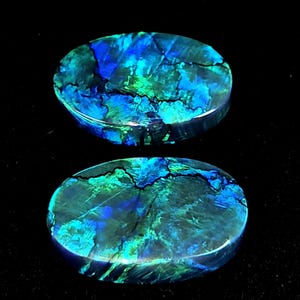 Può includere: Due pietre preziose ovali con una vibrante esposizione di tonalità blu e verdi. Le pietre hanno una superficie lucida con un motivo unico di linee scure, creando un contrasto sorprendente. Le pietre preziose sono su uno sfondo nero.
