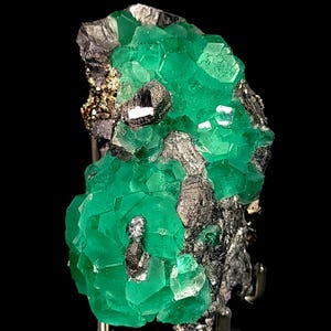 Op de afbeelding: Een levendig groen fluoriet mineraal specimen met een kristallijne structuur. De fluoriet wordt geaccentueerd door donkere, metaalachtig uitziende mineralen, wat een opvallend contrast creëert. Het specimen wordt gepresenteerd tegen een zwarte achtergrond.