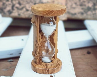 May include: A small wooden hourglass with white sand, sitting on a white wooden surface.