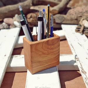 Peut inclure: Un porte-crayons en bois avec une finition en bois naturel. Le porte-crayons est rempli de stylos et de crayons, dont un crayon vert, un stylo noir et un stylo bleu. Le porte-crayons est posé sur une surface en bois blanc.