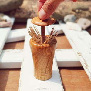 May include: A wooden toothpick holder with a lid. The holder is made of light brown wood with a grain pattern. The lid is a small, round piece of wood with a knob on top. The holder is filled with toothpicks.