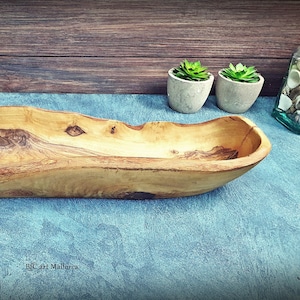 Könnte beinhalten: Eine lange, hölzerne Servierschale mit einer natürlichen, rustikalen Oberfläche. Die Schale ist aus Olivenholz gefertigt und hat eine einzigartige, organische Form.