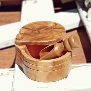 Può includere: Un piccolo saliera rotondo in legno con un coperchio a cerniera e una paletta in legno abbinata. Il saliera è realizzato in legno di ulivo e ha una finitura in legno naturale.