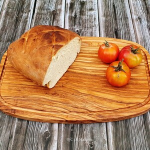 Op de afbeelding: Een houten snijplank met een lichtbruine afwerking en een licht ovale vorm. De plank heeft een verhoogde rand en een groef rondom de omtrek. Een brood en vier rode tomaten liggen op de plank.