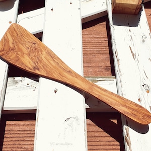 Puede incluir: Una espátula de madera de olivo con una cabeza ancha y plana y un mango largo. La madera muestra un patrón de vetas natural. El fondo es una disposición rústica de tablones de madera pintados de blanco.