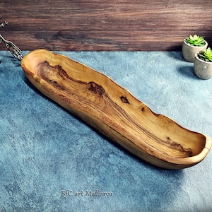 Könnte beinhalten: Eine lange, hölzerne Servierplatte mit einer natürlichen Holzmaserung. Die Platte ist aus einem einzigen Stück Holz geschnitzt und hat ein rustikales, organisches Aussehen. Sie ist perfekt zum Servieren von Brot, Käse oder anderen Vorspeisen.