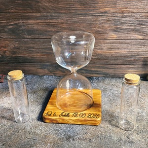 May include: A wooden hourglass stand with a clear glass hourglass and two clear glass vials with cork tops. The stand is engraved with the text "Matt & Julie 12.09.2025".