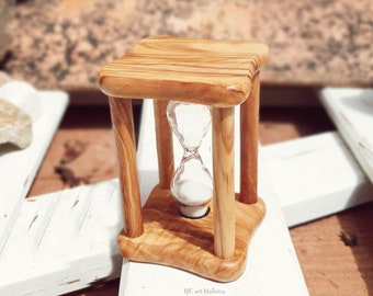 May include: A small wooden hourglass with a white sand timer inside. The hourglass is sitting on a white wooden surface.