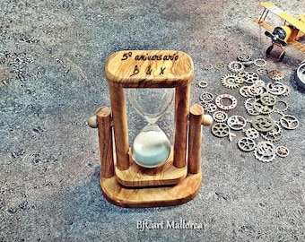 May include: A wooden hourglass with the inscription "5º aniversario B&X" sits on a gray surface surrounded by small metal gears, a compass, and a yellow toy airplane.