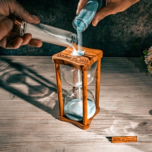 May include: A wooden hourglass with a blue sand timer, engraved with the date "October 05, 2025". The hourglass is being filled with blue sand from a small glass vial.