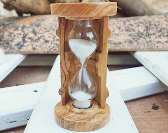 May include: A wooden hourglass with white sand, sitting on a white wooden surface. The hourglass is made of olive wood and has a natural wood grain.