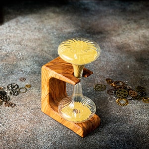 Könnte beinhalten: Eine Sanduhr aus Holz mit einer klaren Glasbirne, die mit gelbem Sand gefüllt ist. Die Sanduhr steht auf einem Holzsockel mit einem dekorativen Design. Der Sockel steht auf einer grauen, strukturierten Oberfläche mit kleinen Metallrädern, die um ihn herum verstreut sind.