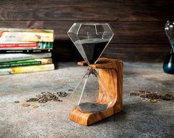 May include: A diamond-shaped hourglass with black sand sits on a wooden stand. The hourglass is surrounded by small metal gears on a gray surface.