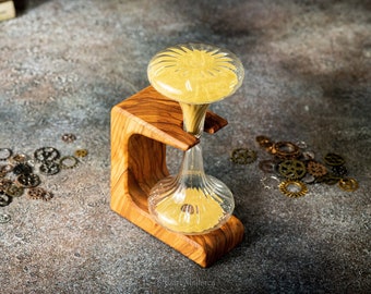 May include: A wooden hourglass with a clear glass bulb filled with yellow sand. The hourglass is sitting on a wooden stand with a decorative design. The stand is on a gray textured surface with small metal gears scattered around it.