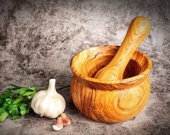 Könnte beinhalten: Ein hölzernes Mörser- und Stößel-Set mit einer Knoblauchzehe, Knoblauchzehen und Petersilienstängeln. Mörser und Stößel sind aus hellem Holz gefertigt. Der Knoblauch ist weiß und die Petersilie grün. Der Hintergrund ist ein gesprenkeltes Grau.