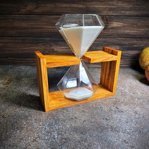 Può includere: Una clessidra a forma di diamante con sabbia bianca poggia su un supporto in legno. La clessidra è in vetro trasparente e ha una base in legno.