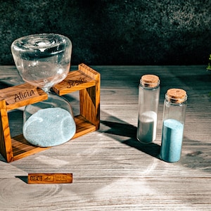 Könnte beinhalten: Eine Sanduhr aus Holz mit den eingravierten Namen "Alicia" und "Jesus". Zwei kleine Glasflaschen mit Korkverschlüssen enthalten blauen und weißen Sand. Die Flaschen stehen auf einer Holzoberfläche mit einem kleinen Holzschild, auf dem "REG Art Handmade" steht.