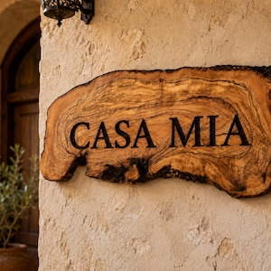 Puede incluir: Un letrero de madera de borde natural con vetas visibles y bordes de corteza oscura, que presenta el texto "CASA MIA" grabado en letras may&uacute;sculas negras. El letrero est&aacute; montado sobre una superficie de pared de piedra clara y texturizada.