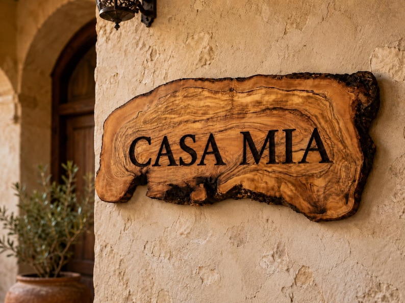 Puede incluir: Un letrero de madera de borde natural con vetas visibles y bordes de corteza oscura, que presenta el texto "CASA MIA" grabado en letras may&uacute;sculas negras. El letrero est&aacute; montado sobre una superficie de pared de piedra clara y texturizada.