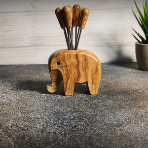 May include: A wooden elephant-shaped appetizer pick holder. The holder is made of light brown wood with a visible grain pattern. It contains several appetizer picks with wooden handles. The elephant has a trunk, ears, and legs. A decorative item for the kitchen.
