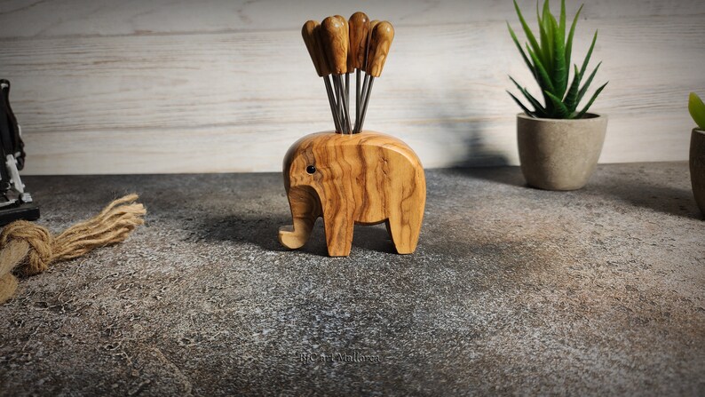 May include: A wooden elephant-shaped appetizer pick holder. The holder is made of light brown wood with a visible grain pattern. It contains several appetizer picks with wooden handles. The elephant has a trunk, ears, and legs. A decorative item for the kitchen.