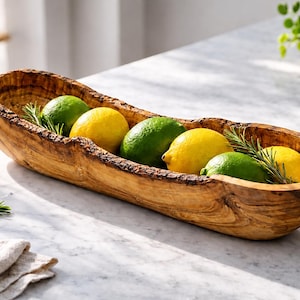 Peut inclure: Un bol en bois rustique rempli de citrons, de citrons verts et de brins de romarin. Le bol est de forme ovale et présente une finition naturelle et texturée. L'arrangement est posé sur un comptoir en marbre.