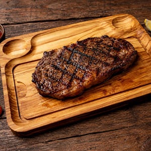 May include: A grilled steak on a wooden serving board with a small bowl of red sauce, lemon wedges, and carrot slices. The steak has grill marks and is surrounded by a wooden serving board with a groove for the sauce.