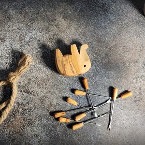 May include: A wooden bunny-shaped fondue pot, with a light brown finish, sits near a set of fondue forks with wooden handles. A decorative rope and small potted plants are also in the image.