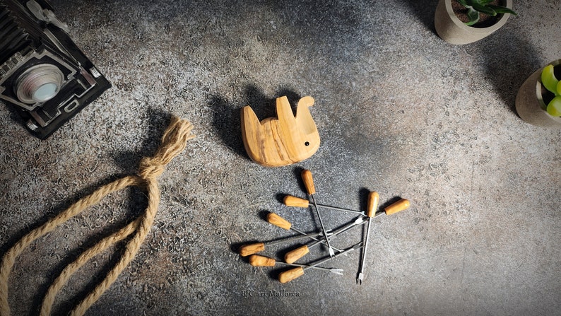May include: A wooden bunny-shaped fondue pot, with a light brown finish, sits near a set of fondue forks with wooden handles. A decorative rope and small potted plants are also in the image.