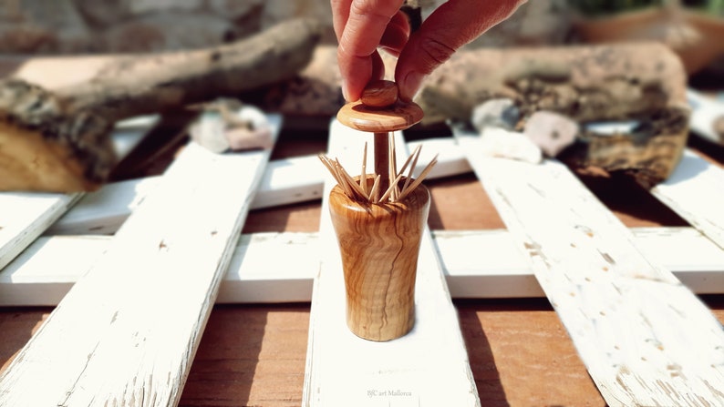 May include: A wooden toothpick holder with a lid shaped like a mushroom. The holder is made of light brown wood with a natural grain pattern. The lid is slightly raised and has a small knob on top. The holder is filled with toothpicks.