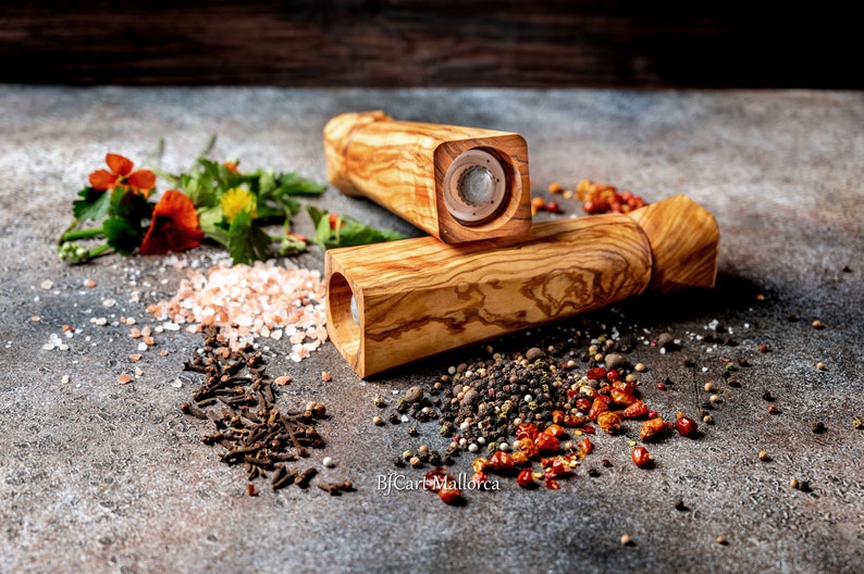 Pu&ograve; includere: Due macinapepe in legno con un motivo a venature naturali sono esposti su una superficie strutturata. Varie spezie, tra cui grani di pepe, chiodi di garofano e peperoncini rossi, sono sparse intorno ai macinapepe. Sono presenti anche erbe fresche e sale rosa.