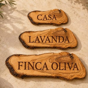 Puede incluir: Tres rodajas de madera de olivo natural con bordes de corteza irregulares, cada una grabada con texto oscuro. La rodaja superior dice CASA, la del medio LAVANDA y la inferior, la m&aacute;s grande, dice FINCA OLIVA. La madera presenta vetas marcadas.