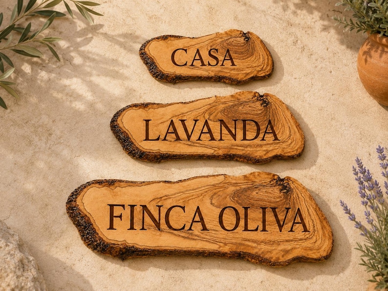 Puede incluir: Tres rodajas de madera de olivo natural con bordes de corteza irregulares, cada una grabada con texto oscuro. La rodaja superior dice CASA, la del medio LAVANDA y la inferior, la m&aacute;s grande, dice FINCA OLIVA. La madera presenta vetas marcadas.
