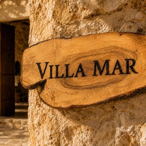 Puede incluir: Un letrero de madera natural con bordes irregulares y rugosos con vetas visibles, que presenta el texto VILLA MAR en letras may&uacute;sculas oscuras y negritas. El letrero est&aacute; montado sobre una superficie de pared de piedra clara y texturizada.