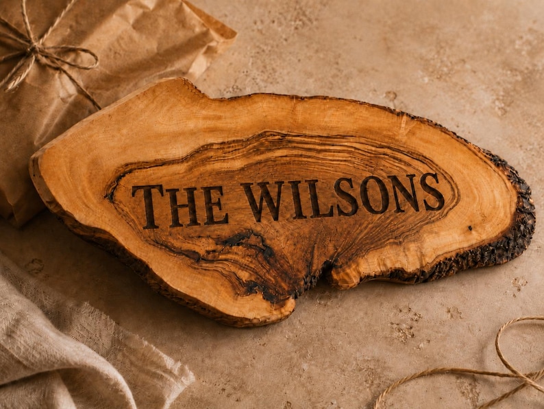 Puede incluir: Una tabla de servir de madera con borde natural, veta marr&oacute;n clara y bordes de corteza oscura. El texto THE WILSONS est&aacute; grabado en una fuente serif oscura en el centro de la tabla, resaltando la forma org&aacute;nica y la textura de la madera.