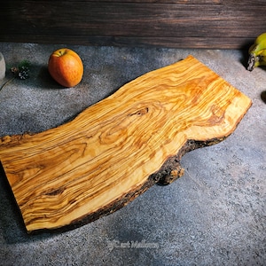 Puede incluir: Una tabla de servir de madera de olivo grande y rústica con un borde natural irregular. La tabla tiene un color marrón intenso con un hermoso patrón de veta.