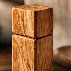 Op de afbeelding: Een houten pepermolen met een vierkant ontwerp. De molen is gemaakt van lichtbruin hout met een opvallend nerfpatroon. Het bovenste gedeelte zit bovenop de basis, waardoor een gestapelde uitstraling ontstaat. De achtergrond is wazig, wat duidt op een keukenomgeving.