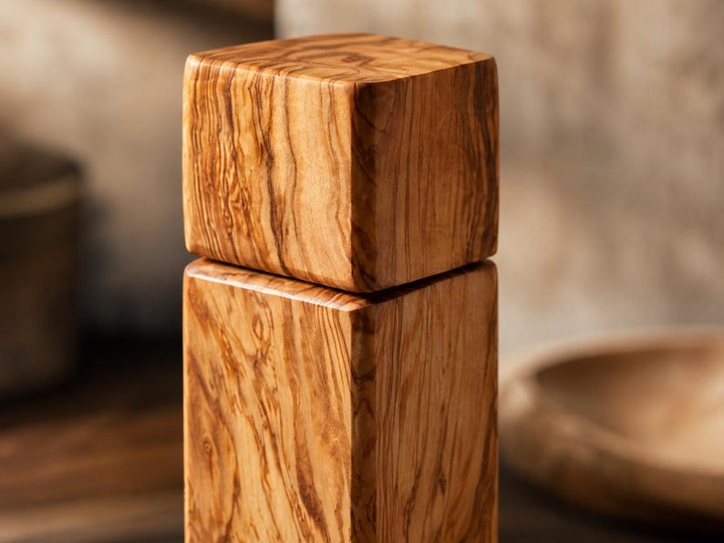 Op de afbeelding: Een houten pepermolen met een vierkant ontwerp. De molen is gemaakt van lichtbruin hout met een opvallend nerfpatroon. Het bovenste gedeelte zit bovenop de basis, waardoor een gestapelde uitstraling ontstaat. De achtergrond is wazig, wat duidt op een keukenomgeving.