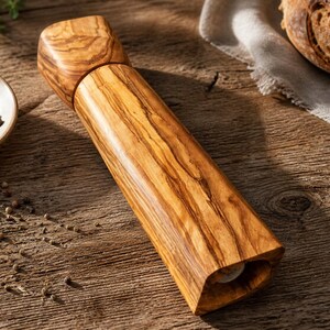 Pu&ograve; includere: Un macinapepe in legno con finitura naturale venata. Il macinino &egrave; cilindrico con parte superiore e inferiore quadrate. A sinistra &egrave; visibile una piccola ciotola di grani di pepe.