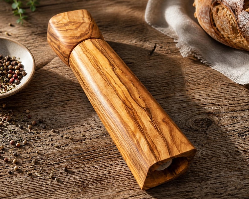 Pu&ograve; includere: Un macinapepe in legno con finitura naturale venata. Il macinino &egrave; cilindrico con parte superiore e inferiore quadrate. A sinistra &egrave; visibile una piccola ciotola di grani di pepe.