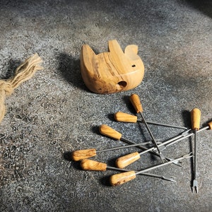 May include: A collection of olive wood fondue forks with wooden handles and a small wooden animal-shaped fondue pot. The forks are stainless steel with a two-pronged end. A vintage camera and rope are also in the image.
