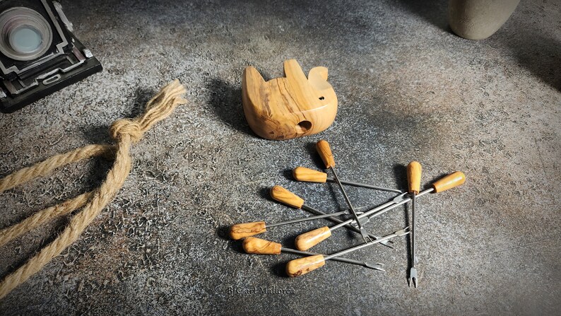 May include: A collection of olive wood fondue forks with wooden handles and a small wooden animal-shaped fondue pot. The forks are stainless steel with a two-pronged end. A vintage camera and rope are also in the image.