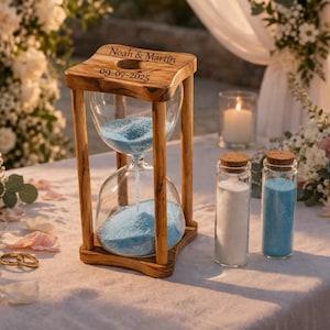 Puede incluir: Un reloj de arena de madera con arena azul, grabado con "Noah & Martin" y la fecha 09-07-2025. Dos pequeñas botellas de vidrio con arena blanca y azul están junto al reloj de arena. La escena está ambientada sobre un mantel blanco con arreglos florales.