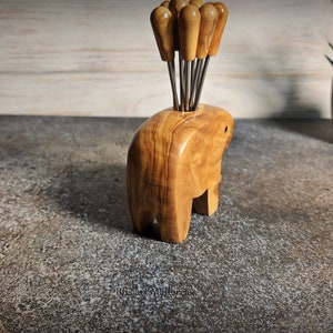 May include: A wooden elephant-shaped toothpick holder with a collection of toothpicks. The holder is made of light brown wood and has a smooth finish. The toothpicks have wooden handles and metal tips. A decorative item for the kitchen or dining room.