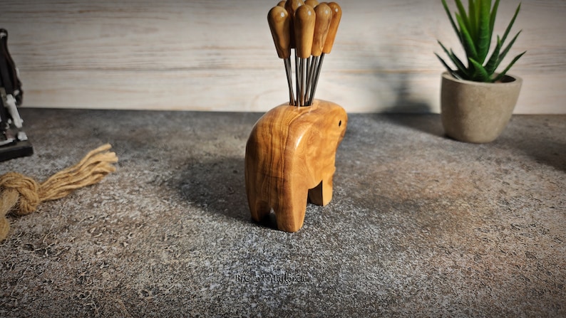 May include: A wooden elephant-shaped toothpick holder with a collection of toothpicks. The holder is made of light brown wood and has a smooth finish. The toothpicks have wooden handles and metal tips. A decorative item for the kitchen or dining room.
