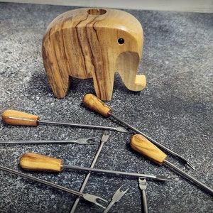 May include: A wooden elephant-shaped candle holder with a small hole on top, accompanied by several fondue forks. The forks have wooden handles and stainless steel tines. The items are arranged on a textured gray surface.