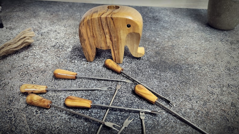 May include: A wooden elephant-shaped candle holder with a small hole on top, accompanied by several fondue forks. The forks have wooden handles and stainless steel tines. The items are arranged on a textured gray surface.
