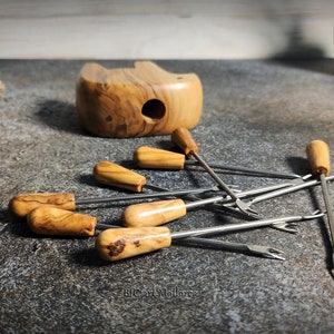 May include: A collection of fondue forks with wooden handles and stainless steel tines, scattered on a textured surface. A wooden bowl and a small plant are in the background. The forks have a classic design, suitable for serving cheese or chocolate fondue.