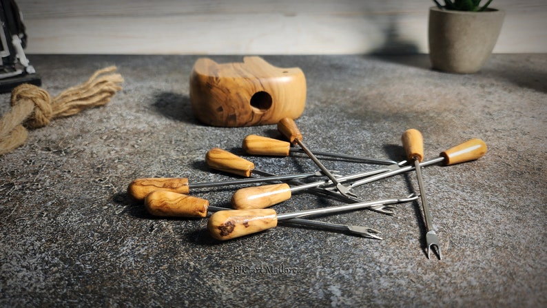 May include: A collection of fondue forks with wooden handles and stainless steel tines, scattered on a textured surface. A wooden bowl and a small plant are in the background. The forks have a classic design, suitable for serving cheese or chocolate fondue.