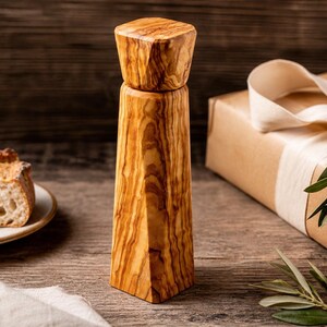 Pu&ograve; includere: Un macinapepe in legno dal design geometrico, che mostra la venatura naturale del legno. Nell'immagine sono presenti anche un piatto con fette di pane e un regalo avvolto con un nastro e del verde. Il macinapepe &egrave; alto circa 18 cm.